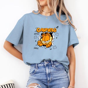 May include: Light blue t-shirt featuring the cartoon character Garfield bursting through a wall. The word "GARFIELD" is printed in orange and black above the character. Additional Garfield graphics are on the shirt.