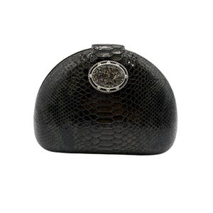 Puede incluir: Un bolso de mano negro en forma de semicírculo con una textura brillante similar a la piel de serpiente. La parte frontal presenta un cierre metálico ovalado central con una piedra cristalina oscura y texturizada, rodeada por un borde plateado.