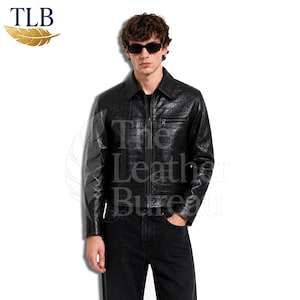 Peut inclure: Une veste noire avec un motif texturé en relief façon crocodile, dotée d'un col, d'une fermeture éclair intégrale sur le devant et d'une poche poitrine. Le vêtement est porté avec un pantalon noir et des lunettes de soleil. Texte TLB et The Leather Bureau.