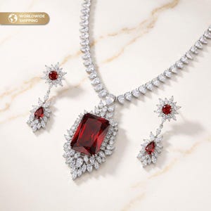May include: A dazzling jewelry set featuring a necklace and earrings. The necklace has a large, rectangular, deep red gemstone pendant surrounded by white stones. The earrings have a similar design with red and white stones. The set is on a marble surface.