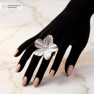 Könnte beinhalten: Ein silberfarbener Blumenring mit detailliertem Blütenblattdesign. Der Ring wird auf einem schwarzen Handschuh getragen, mit dem Text "WORLDWIDE SHIPPING" oben links. Die Blütenblätter des Rings haben ein strukturiertes Aussehen.