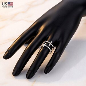 May include: A silver-colored ring with a double-band design, featuring two cross motifs. The top cross is embellished with small, clear stones. The ring is displayed on a black hand against a marble-patterned surface.