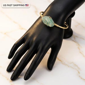 May include: A gold-colored bracelet featuring a raw, light green gemstone wrapped in wire. The bracelet is displayed on a black hand against a marble background. The bracelet's design is simple and elegant.