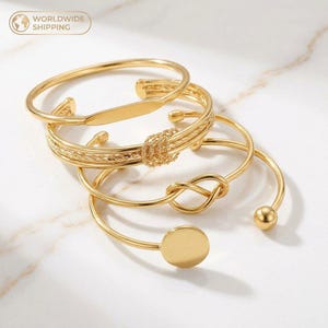May include: Four gold-coloured bangles in various designs. One has a rectangular plate, another features a woven knot, a third has a knot design, and the last has a small round disc. The bracelets are displayed on a white surface.