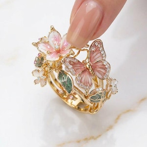 May include: A gold ring with a butterfly and floral design. The ring features pink and white enamel flowers, a pink butterfly with crystal accents, and green leaves. The ring is held up by a finger.