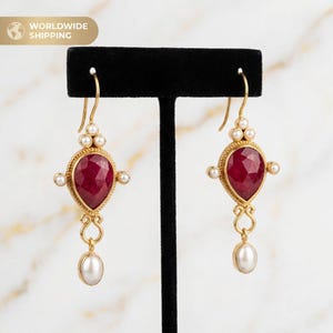 Puede incluir: Pendientes dorados con un centro de piedra preciosa roja oscura en forma de lágrima, rodeado por un marco dorado y pequeñas perlas. Los pendientes tienen una perla colgante en la parte inferior y se exhiben sobre un soporte negro. El texto "WORLDWIDE SHIPPING" está en la esquina superior izquierda.