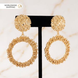 Può includere: Orecchini a cerchio dorati con un design strutturato a forma di fiore. Gli orecchini presentano un design a cerchio circolare sospeso da un perno rotondo. Sono esposti su un supporto nero su sfondo chiaro. Il testo "WORLDWIDE SHIPPING" è visibile.
