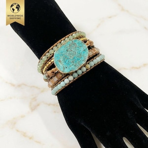 Op de afbeelding: Een multi-strand wikkelarmband met een grote, ovale turkoois steen. De armband bevat groene, bruine en blauwe kralen, met gouden accenten. De armband wordt gepresenteerd op een zwart oppervlak. Ongeveer 19 cm lang.
