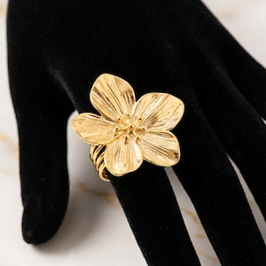 May include: A gold-colored flower ring with detailed petal design. The ring is displayed on a black surface, showcasing the intricate details of the floral design. The ring's band has a textured, layered appearance.