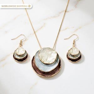 Könnte beinhalten: Ein goldfarbenes Schmuckset mit einer Anhänger-Halskette und passenden Ohrringen. Der Anhänger und die Ohrringe haben ein geschichtetes, kreisförmiges Design mit weißem, hellblauem und braunem Emaille. Auf einer weißen Marmoroberfläche.