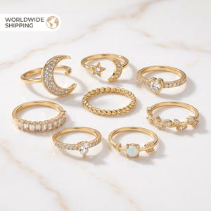 May include: A collection of gold-toned rings with various designs, including a crescent moon, star, heart, and leaf motifs. Some rings feature clear gemstones, while others have a textured or braided appearance. The rings are displayed on a white marble surface.