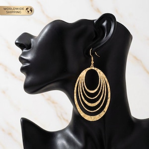 May include: Gold-toned oval earrings with a textured finish. The earrings feature multiple nested oval shapes, creating a layered effect. The earrings are displayed on a black mannequin head against a marble background.