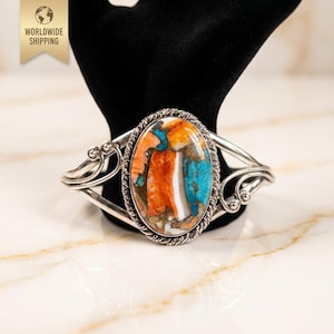 May include: A silver cuff bracelet with an oval-shaped, multicolored stone. The stone features orange, turquoise, white, and brown hues. The bracelet has a twisted rope design around the stone and decorative swirls on the sides. The image also includes the text "WORLDWIDE SHIPPING".