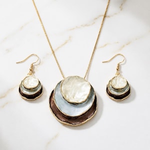 May include: A gold-tone necklace and earring set featuring layered, circular pendants. The necklace has a thin chain and three stacked discs in white, light blue, and dark brown. The matching earrings mirror this design with smaller stacked discs.