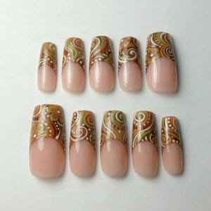 Puede incluir: Un juego de diez uñas postizas con diseño de manicura francesa, base color nude y patrones complejos de flores y espirales en marrón, verde oliva y blanco. Las uñas tienen un acabado brillante y están dispuestas en dos filas de cinco.