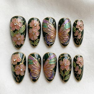 May include: A set of ten almond-shaped press-on nails with a dark green base. The designs feature hand-painted pink flowers with small clear rhinestones at the centers and detailed butterfly wings in shades of purple, green, and pink with gold outlines.
