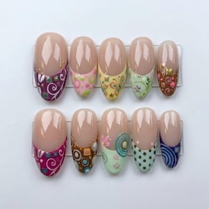 Puede incluir: Un juego de diez uñas postizas ovaladas con base color nude y puntas decoradas. Los diseños incluyen espirales moradas, círculos rosas, mariposas amarillas, motivos florales verdes, cuadrados marrones, puntos y círculos azules.