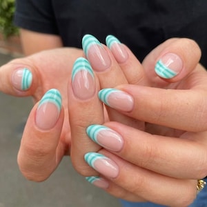 Puede incluir: Uñas de forma ovalada con estilo de manicura francesa, con una base de color nude y puntas con rayas horizontales en color turquesa claro y blanco. El diseño de uñas cubre el borde libre de cada uña, creando un patrón de rayas limpio.