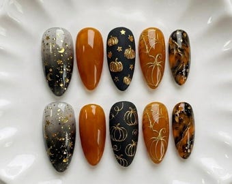 May include: A set of ten almond-shaped press-on nails featuring various designs including black-to-clear gradients with gold stars and moons, solid amber, matte black with gold pumpkins, amber with gold spiderwebs, and black and amber tortoiseshell patterns.