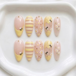 May include: A set of ten almond-shaped press-on nails featuring a nude base with various designs including yellow French tips with bees, yellow horizontal stripes, yellow and brown polka dots, yellow honeycomb patterns with bees, and white daisies.