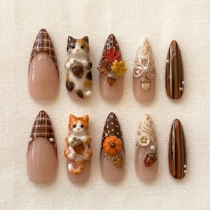 May include: A set of ten almond-shaped press-on nails featuring autumn-themed designs. The collection includes brown plaid patterns, three-dimensional cat figures holding acorns, maple leaves, pumpkins, white swirls, buttons, and vertical brown stripes.