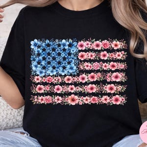 May include: Black t-shirt with a floral American flag design. The flag's stars are made of blue flowers, and the stripes are made of pink flowers. The shirt is a casual style.