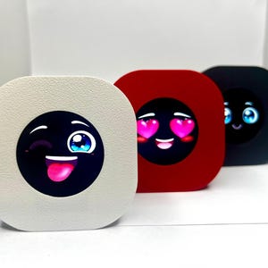 May include: Three square devices in a row, each in a different colour: off-white, red, and black. Each device has a circular screen displaying a different emoji face. The off-white device has a winking face with its tongue out. The red device has a face with heart eyes. The black device has a smiling face.