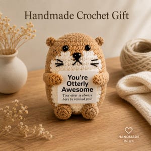 May include: A handmade crochet otter figurine in shades of brown and cream, holding a small sign that reads "You're Otterly Awesome." The text "Handmade Crochet Gift" is at the top. The figurine has black eyes and whiskers.