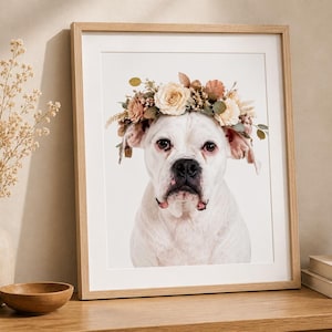 May include: A framed wall art print featuring a portrait of a white dog wearing a floral crown made of cream, pink, and brown flowers and green leaves. The dog has a black nose and dark eyes. The frame is light-colored wood with a white mat border.
