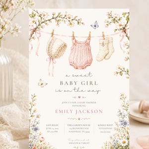 May include: A baby shower invitation card featuring a watercolor illustration of baby clothes hanging on a clothesline, including a bonnet, romper, and socks, surrounded by floral branches, butterflies, and the text A sweet baby girl is on the way.