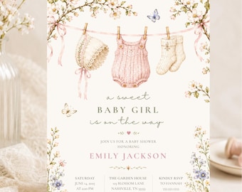 Puede incluir: Una tarjeta de invitación para baby shower con una ilustración en acuarela de ropa de bebé colgada en un tendedero, incluyendo un gorrito, un mameluco y calcetines, rodeados de ramas florales, mariposas y el texto A sweet baby girl is on the way.