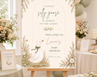 Puede incluir: Un letrero rectangular de bienvenida para baby shower sobre un caballete de madera con una oca blanca con cinta verde, detalles florales y el texto A LITTLE silly goose IS ALMOST ON THE LOOSE! WELCOME TO Lucia's BABY SHOWER JUNE 22, 2025.