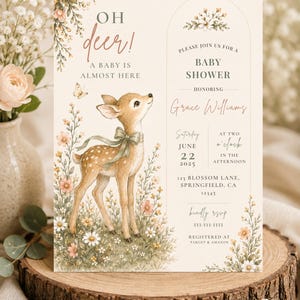 May include: A cream-colored baby shower invitation card featuring a watercolor illustration of a fawn with a green ribbon, surrounded by wildflowers and a butterfly. The text reads: Oh deer! A baby is almost here, Please join us for a baby shower honoring Grace Williams, Saturday June 22 2025 at two o'clock, 123 Blossom Lane, Springfield, CA 12345, kindly RSVP 333-333-3333, registered at Target & Amazon.