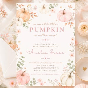 May include: A rectangular baby shower invitation card featuring watercolor illustrations of pink and white pumpkins, green eucalyptus leaves, and small pink flowers. The text reads: a sweet little PUMPKIN is on the way! PLEASE JOIN US FOR A BABY SHOWER HONORING Amelia Grace SATURDAY, OCTOBER 13TH AT 2:00 PM THE PUMPKIN PATCH 1234 FALL GROVE, ORLANDO, FL RSVP TO JANE AT 555-555-5555 REGISTERED AT AMAZON & TARGET.