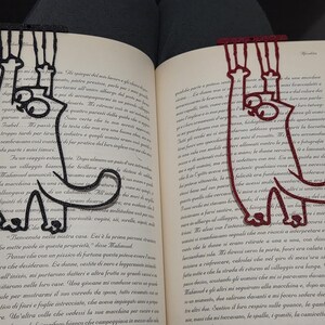 Può includere: Due segnalibri a forma di gatto sono posizionati in un libro aperto. Uno è nero, l'altro bordeaux. I segnalibri sono realizzati in un materiale sottile e flessibile e hanno un design stravagante. Il libro è aperto su una pagina di testo.