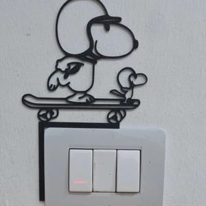 Può includere: Copri interruttore Snoopy in metallo nero. Il design presenta Snoopy su uno skateboard sopra un interruttore della luce con tre pulsanti bianchi. L'interruttore è fissato a una parete bianca.