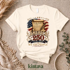 Puede incluir: Camiseta color crema con un diseño patriótico que celebra el 250 aniversario de América. El gráfico incluye la bandera estadounidense, un pergamino con el texto "We the People" y los años 1776-2026. La camiseta tiene mangas cortas.