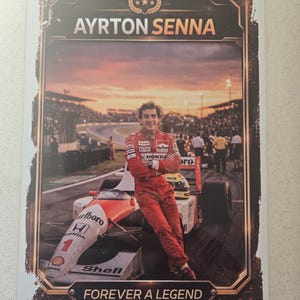 Puede incluir: Un póster con Ayrton Senna con un traje de carreras rojo, apoyado en un coche de Fórmula 1. El póster incluye el texto "AYRTON SENNA" y "FOREVER A LEGEND" con un fondo de atardecer y una pista de carreras.
