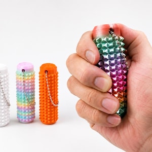 May include: Four cylindrical sensory fidget tools with a textured, spiked surface. One is solid white, one is solid orange, one features a pastel rainbow gradient, and one has a vibrant rainbow gradient, each equipped with a small metal ball chain.