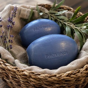 Pode incluir: Dois sabonetes ovais de cor azul com a palavra PROVENDI gravada em cada um, colocados numa cesta de vime forrada com um pano de linho bege, acompanhados por ramos de lavanda roxa seca e folhas de oliveira verdes.
