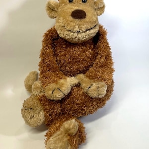 Pode incluir: Um peluche de macaco com pelo castanho felpudo no corpo e pelo bege liso no rosto, orelhas, mãos e pés. O animal de peluche apresenta olhos de plástico preto, um nariz castanho escuro e uma boca costurada, em uma posição sentada.