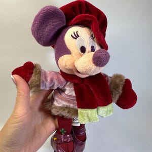 May include: A plush Minnie Mouse doll wearing a deep red velvet hat, a matching red scarf with light green ends, and a light pink velvet jacket with brown faux fur trim. The doll has dark red mittens, dark red leggings, and dark red shoes.