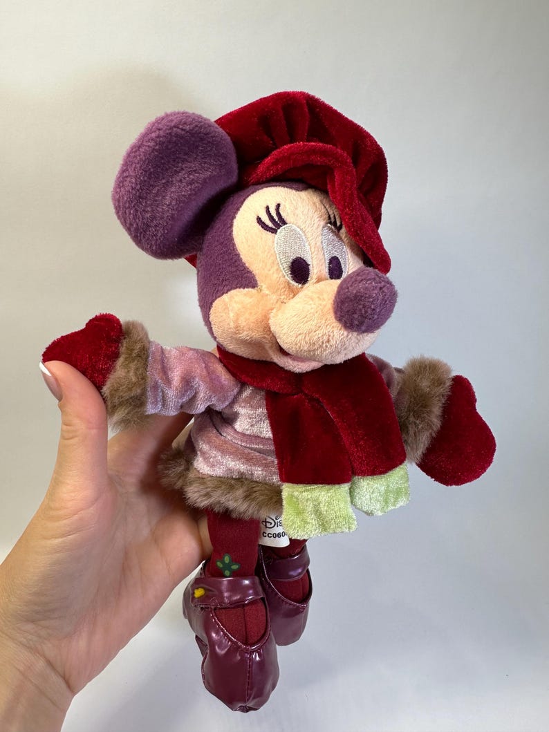 May include: A plush Minnie Mouse doll wearing a deep red velvet hat, a matching red scarf with light green ends, and a light pink velvet jacket with brown faux fur trim. The doll has dark red mittens, dark red leggings, and dark red shoes.