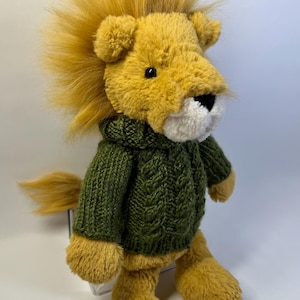Puede incluir: Un león de peluche con pelaje amarillo dorado, una melena esponjosa y un hocico blanco. El león lleva puesto un suéter de punto verde oliva oscuro con un patrón de trenzas verticales en la parte delantera, mangas largas y cuello alto.
