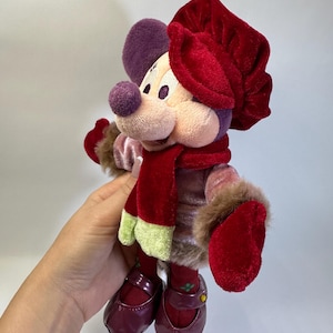 May include: A plush mouse character wearing a red velvet hat, a matching red scarf with light green ends, and a pink velvet jacket with brown faux fur trim at the cuffs. The character has dark red shoes and purple ears, presented in a soft toy form.