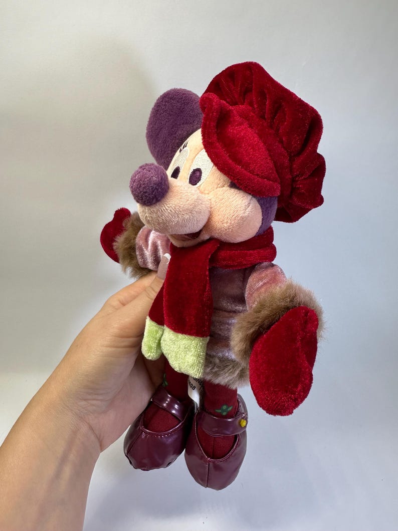 May include: A plush mouse character wearing a red velvet hat, a matching red scarf with light green ends, and a pink velvet jacket with brown faux fur trim at the cuffs. The character has dark red shoes and purple ears, presented in a soft toy form.