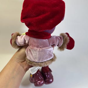 May include: A plush doll wearing a deep red velvet hat, a matching red scarf, and red mittens. The doll is dressed in a light pink velvet coat with brown faux fur trim at the cuffs and hem, paired with dark red shoes with small yellow accents.