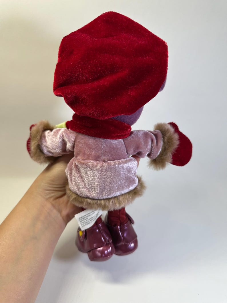 May include: A plush doll wearing a deep red velvet hat, a matching red scarf, and red mittens. The doll is dressed in a light pink velvet coat with brown faux fur trim at the cuffs and hem, paired with dark red shoes with small yellow accents.