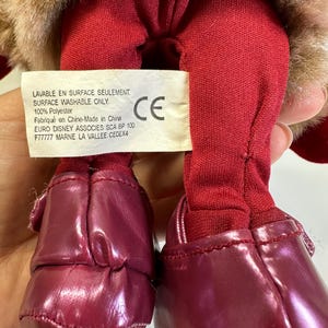 May include: A plush toy with brown faux fur, red fabric legs, and shiny dark red shoes. A white rectangular tag is attached, featuring the text: LAVABLE EN SURFACE SEULEMENT, SURFACE WASHABLE ONLY, 100% Polyester, Fabriqu&eacute; en Chine-Made in China, EURO DISNEY ASSOCIES SCA BP 100 F77777 MARNE LA VALLEE CEDEX4.