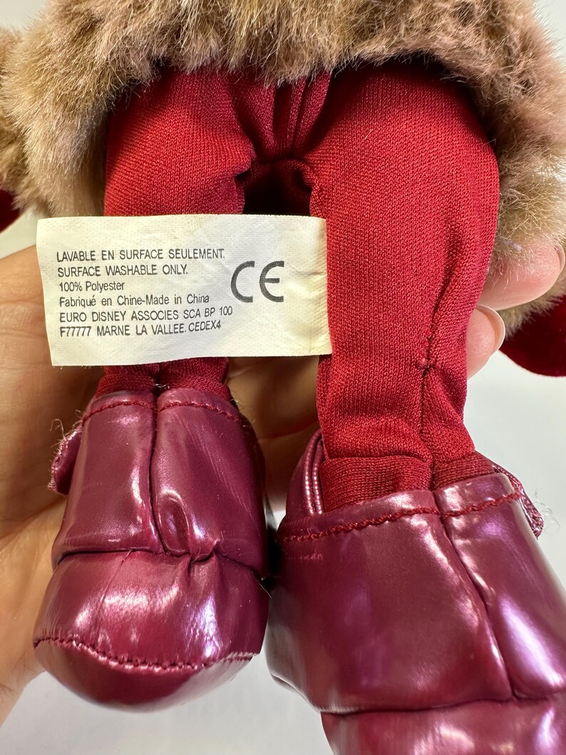May include: A plush toy with brown faux fur, red fabric legs, and shiny dark red shoes. A white rectangular tag is attached, featuring the text: LAVABLE EN SURFACE SEULEMENT, SURFACE WASHABLE ONLY, 100% Polyester, Fabriqu&eacute; en Chine-Made in China, EURO DISNEY ASSOCIES SCA BP 100 F77777 MARNE LA VALLEE CEDEX4.